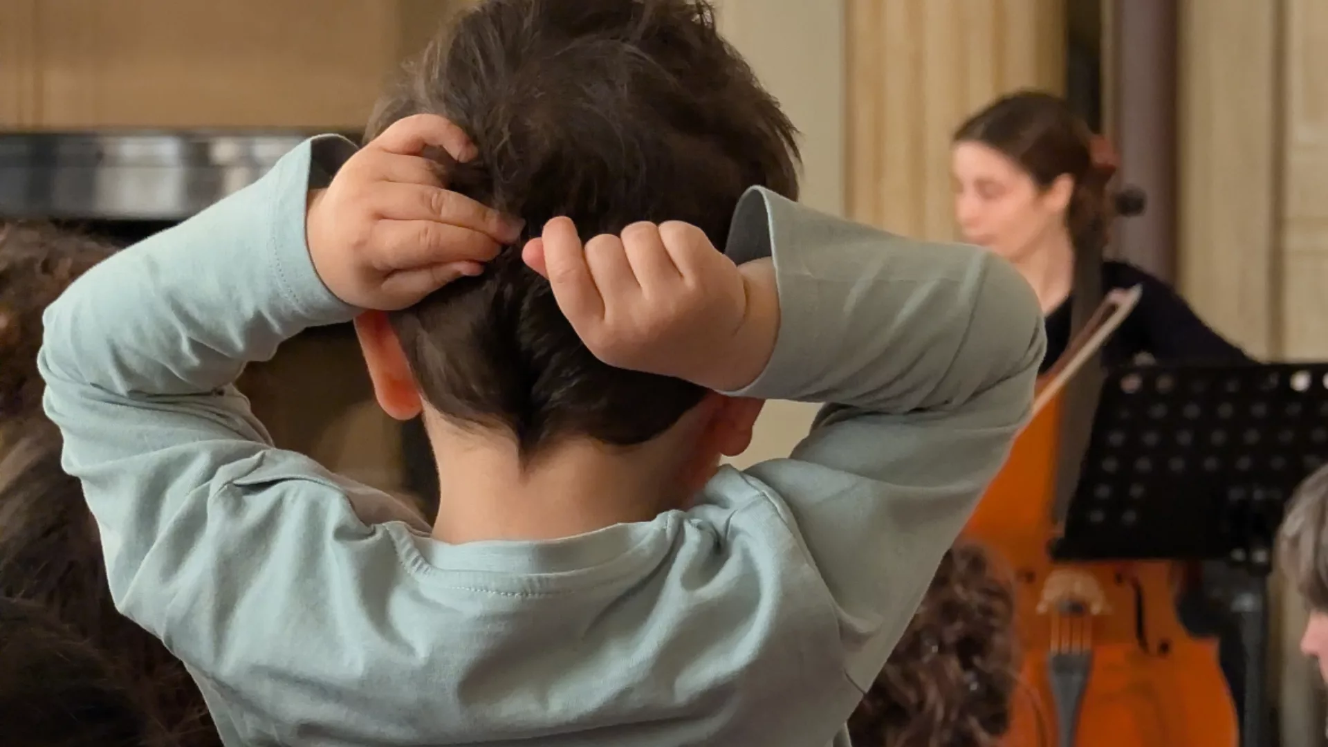 Un bambino visto di spalle che si accarezza i capelli mentre guarda una violoncellista che suona